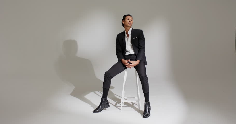 Asian male model shifting, standing, draping suit jacket on stool under studio spotlight for shoot. Fashion, elegance, professionalism, portrait, minimalism, male, studio