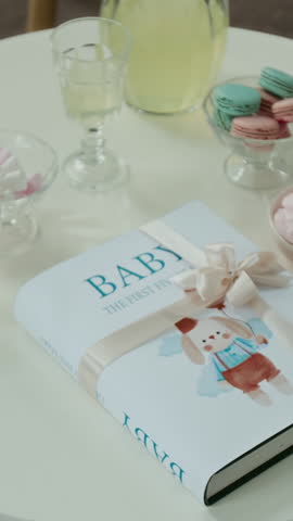 No people vertical close-up of baby care book with pink ribbon on table, gifted to pregnant woman by friend or guest at gender reveal or baby shower party, next to pink, blue desserts, jug of lemonade