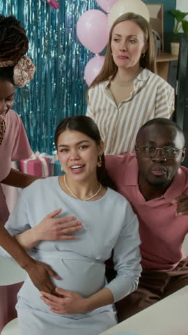 Medium vertical portrait of beautiful young pregnant Hispanic woman, African American husband and diverse girlfriends posing for photos at gender reveal party, smiling, waving, blowing kisses