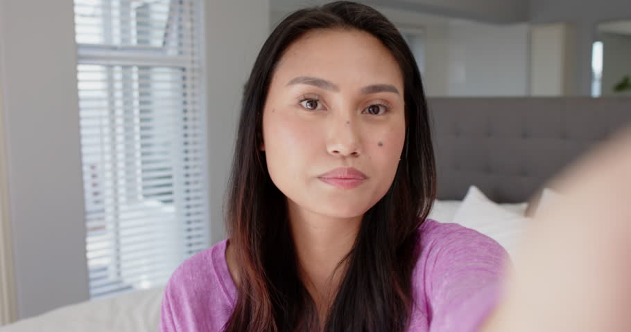 Mid adult Asian woman vlogging update with smartphone, adjusting camera, laughing, speaking on bed. Vlogging, lifestyle, casual, modern, comfort, authenticity, communication