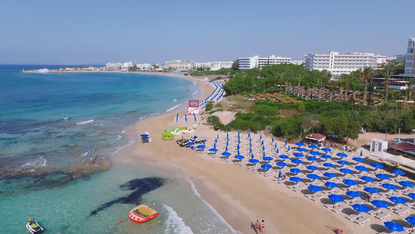 A slow pan of Ayia Napa beach in Cyprus, showing golden sand, turquoise waters, sunbeds, a lifeguard tower, and resort buildings with greenery.