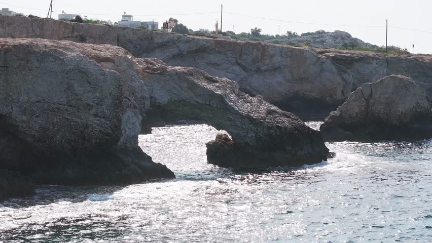 Rugged cliffs and clear blue waters in Ayia Napa, Cyprus. Gentle waves lap against the shore with palm trees and white buildings in the background.