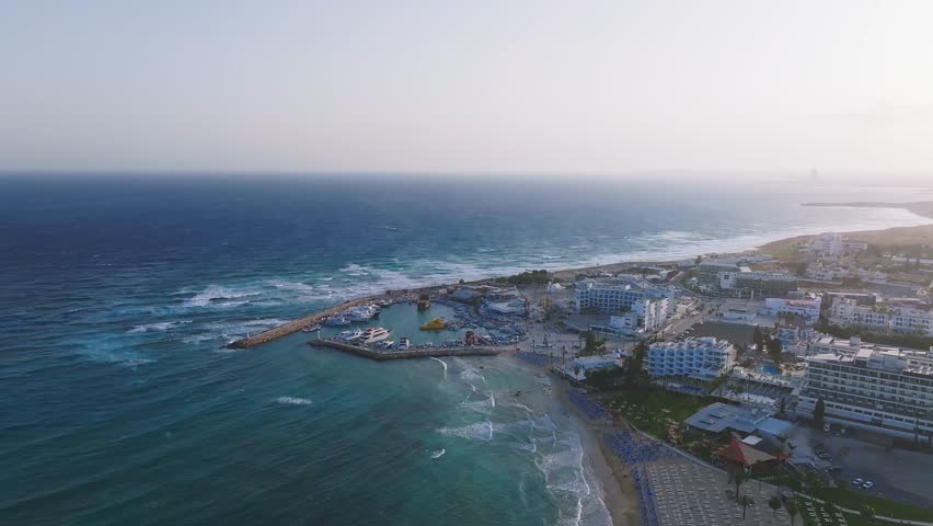 Aerial sweep over Ayia Napa, Cyprus, showcasing the coastline, sandy beaches, marina, modern resorts, and town layout during sunset.