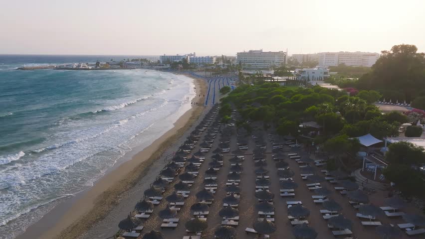 Aerial view of a beach in Ayia Napa, Cyprus, with blue sun loungers, turquoise waves, resort buildings, and palm trees under a warm sunlit sky.