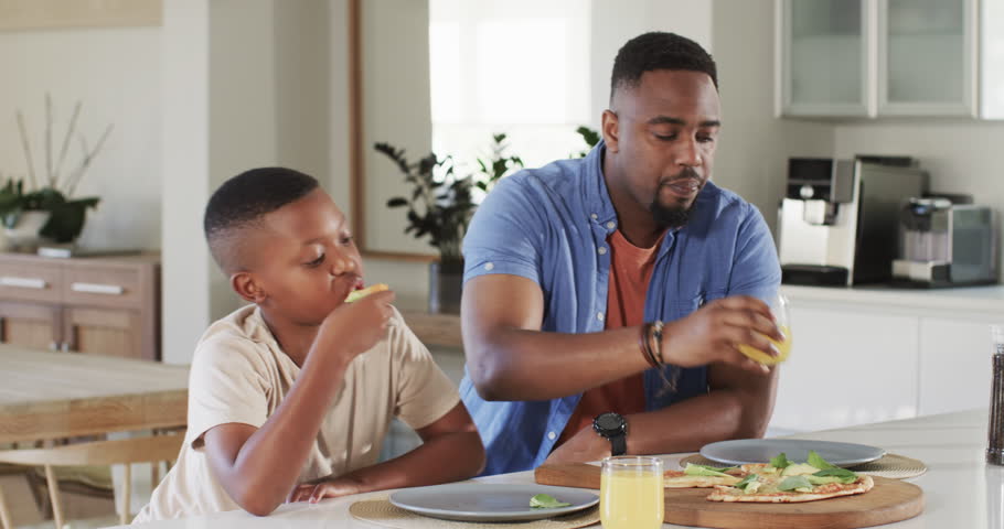 African American father and son picking pizza, pouring juice, clinking glasses at kitchen island. Family, bonding, togetherness, culinary, modern, wholesome, cheerful