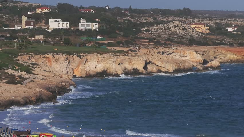 Aerial view of Ayia Napa, Cyprus, showing vibrant blue waters, rugged orange cliffs, whitewashed buildings, and gentle waves along the shoreline.