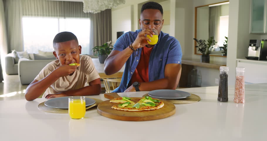 African American father and son in kitchen after boy biting pizza father clinking juice son eating. Family, bonding, domestic, nutrition, togetherness, lifestyle, warmth