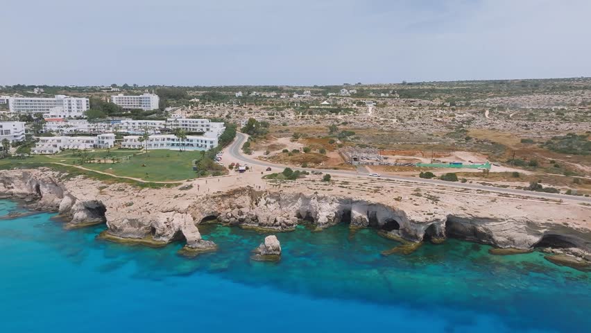 Aerial pan of Ayia Napa, Cyprus, showing resorts on rocky cliffs, turquoise Mediterranean waters, sea caves, and sparse vegetation.