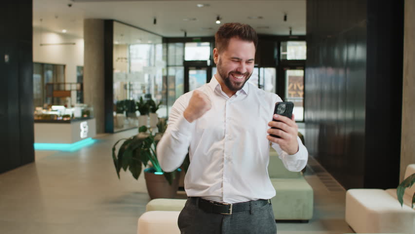 Happy young businessman guy in white shirt working on smartphone shocked by sudden victory game winning lottery jackpot achievement good career news. Excited man in formal standing in office lobby.