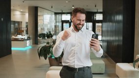 Happy young businessman guy in white shirt working on smartphone shocked by sudden victory game winning lottery jackpot achievement good career news. Excited man in formal standing in office lobby. - Powered by Shutterstock - Get 15% off with code: PIKWIZARD15
