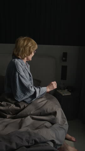 Vertical side view shot of senior woman taking pills in morning and drinking glass of water while sitting on bed
