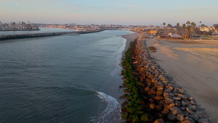 Aerial flyover of the San Gabriel River at golden hour with waves crashing into the rocky jetties, Seal Beach, California