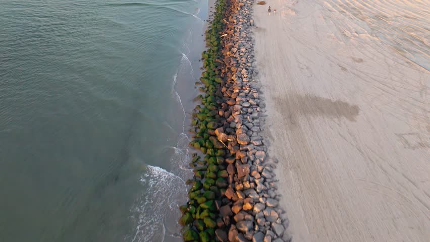 Aerial flyover of the San Gabriel River at golden hour with waves crashing into the rocky jetties, Seal Beach, California