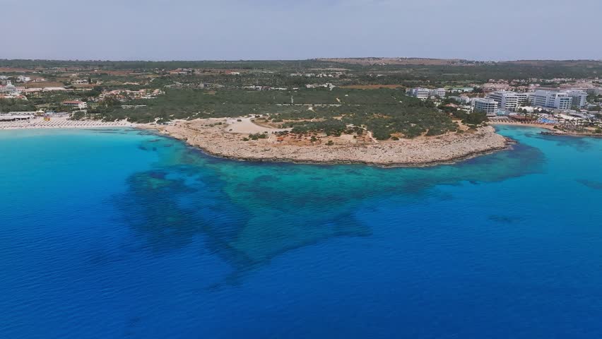 Aerial view of Ayia Napa, Cyprus, showing turquoise waters, rocky shores, beach umbrellas, parked vehicles, and resorts amid sandy terrain.