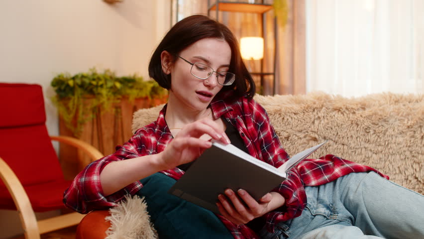 Young woman sits on home sofa, reading through old personal journal notebook and smiling with nostalgic mood. Girl reflects on memories with calm expression, embracing past thoughts and quiet emotion.