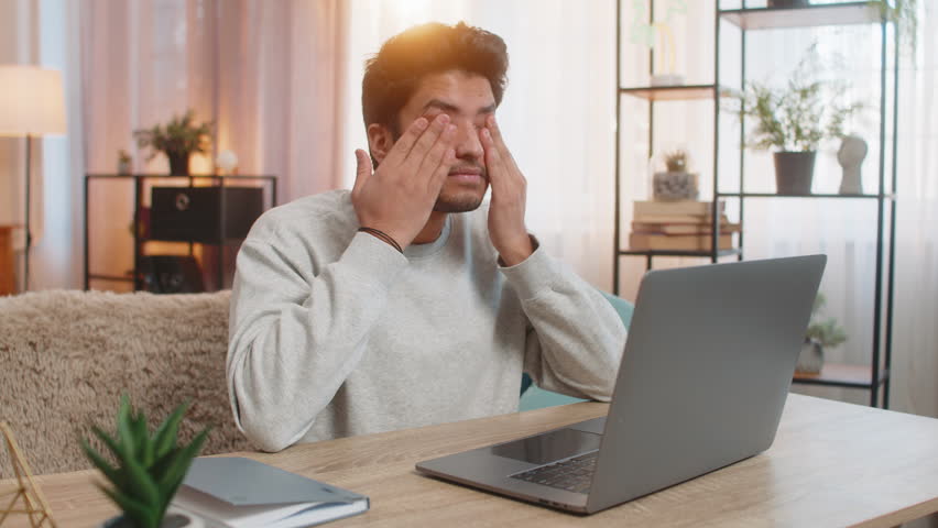 Young Indian man freelancer working on laptop, looking tired, rubbing eyes while sitting at table on sofa in living room at home. Hispanic guy, a workaholic, continues his tasks, clearly exhausted.