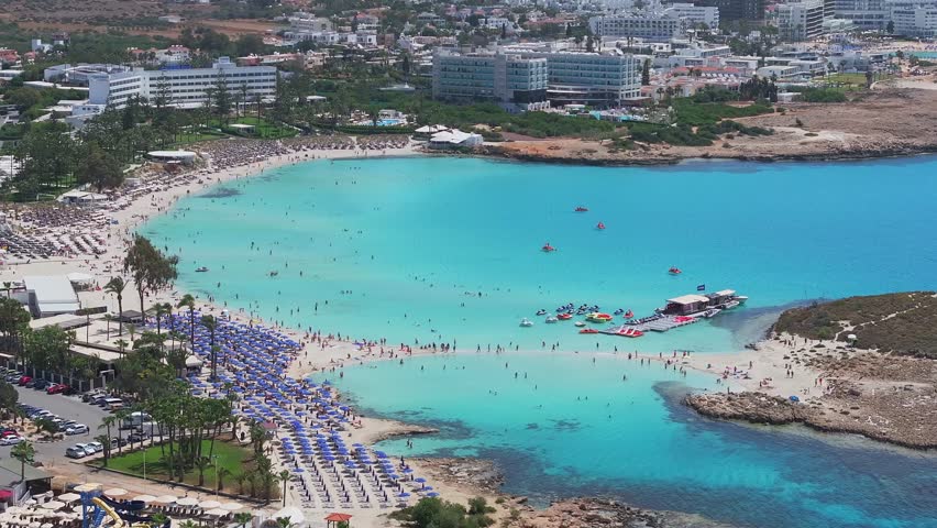Aerial view of Ayia Napa, Cyprus, showing turquoise waters, a sandy Nissi beach with a rocky island, a docked boat, and a speedboat in motion.
