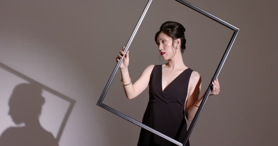 Spotlight casting shadow guiding Asian woman sliding large frame diagonally in studio, copy space. Silhouette, minimalism, elegance, composition, artistic, fashion, contrast