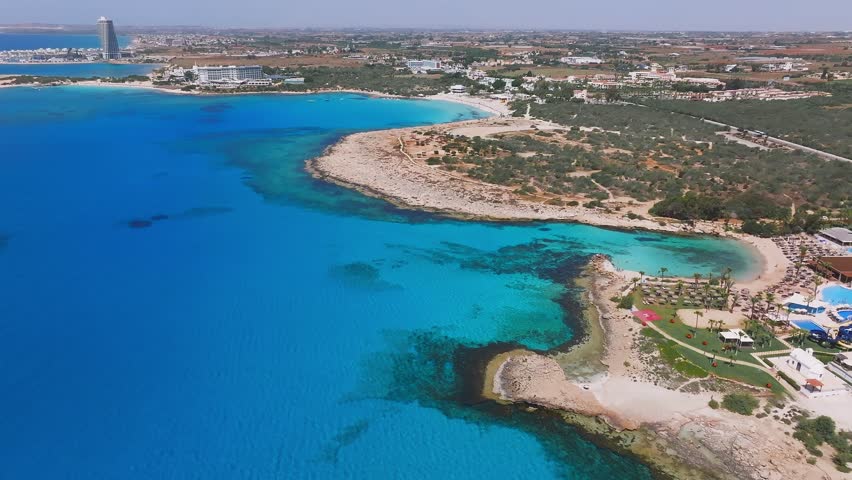 Aerial view of Ayia Napa, Cyprus, showing turquoise waters, rocky shores, Love Arch, resorts, Nissi Beach, Landa Beach, and gentle wave motion.