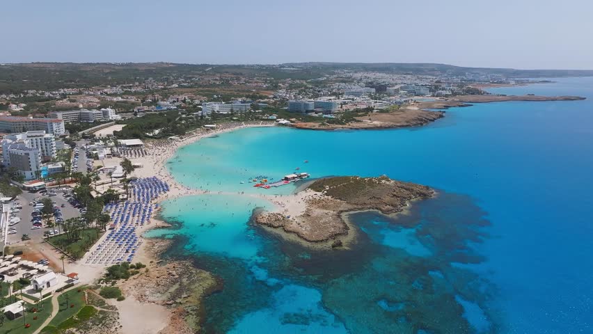 Aerial view of Ayia Napa, Cyprus, showcasing Nissi Beach, turquoise waters, modern resorts, rocky outcrops, boats, and vibrant beach activity.