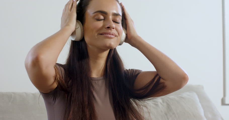 Woman placing white headphones, tapping phone, selecting music to relax, reclining on home sofa. Relaxation, solitude, comfort, leisure, tranquility, lifestyle, modern