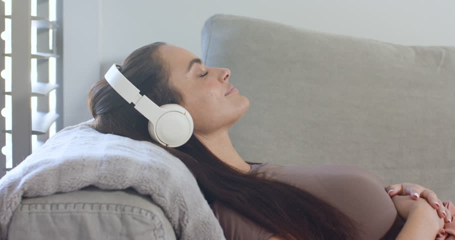 Woman placing white headphones, tapping phone, selecting music to relax, reclining on home sofa. Relaxation, solitude, comfort, leisure, tranquility, lifestyle, modern