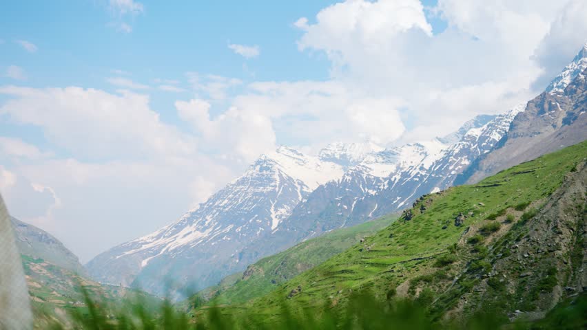 4K shot of fluffy clouds above the snowy Himalayan mountain peaks during the summer season with defocused green grass moving due to strong winds at Sissu village in Lahaul, Himachal Pradesh, India.