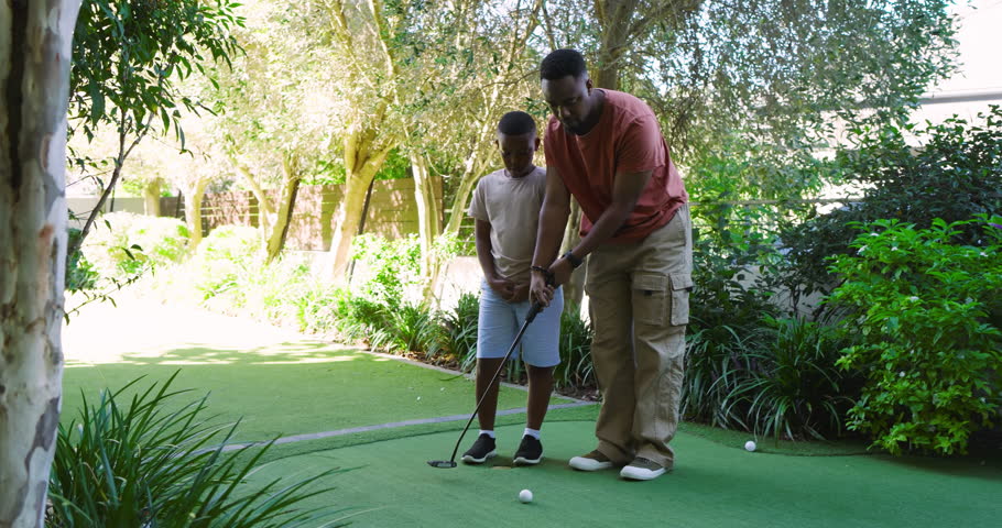 African American father and son at backyard green guiding putter grip, ball dropping, teaching golf. Mentorship, family, outdoor, leisure, recreation, bonding, natural