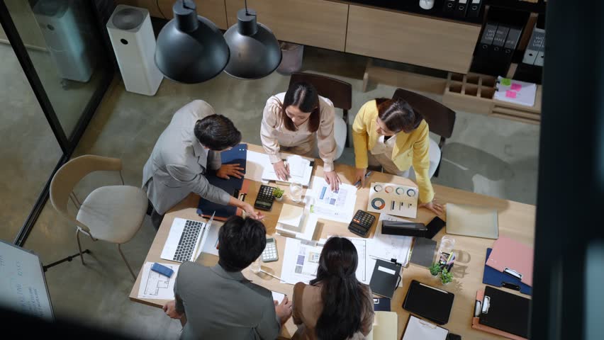 High angle of diverse business team engaged in strategy planning session in modern home office space, using laptops and reviewing printed business documents
