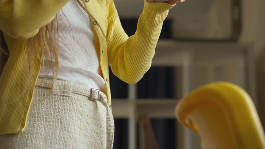 A close-up shows a person wearing a sunny yellow cardigan, a white top, and a patterned skirt, holding an indiscernible object. The indoor setting is cozy, with a bookshelf visible in the backdrop.