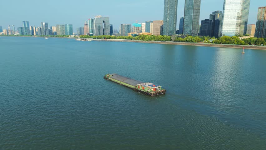 Aerial view of barge navigates river in urban setting with modern skyscrapers during clear day. Hangzhou, China.