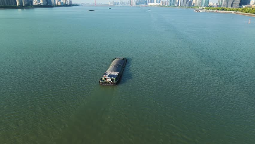 Aerial view of barge navigates river in urban setting with modern skyscrapers during clear day. Hangzhou, China.