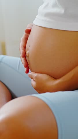 Asian Close up of pregnant woman gently holding her belly, wearing white top and light blue pants, symbolizing care and anticipation during pregnancy and motherhood. Vertical Video