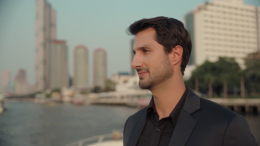 Portrait of confident Latin American businessman in suit smiles to camera on yacht deck. Happy Hispanic man enjoys relaxing trip on modern motorboat against downtown skyscrapers