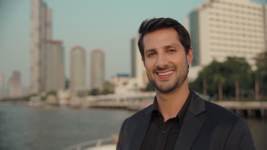 Portrait of confident Latin American businessman in suit smiles to camera on yacht deck. Happy Hispanic man enjoys relaxing trip on modern motorboat against downtown skyscrapers