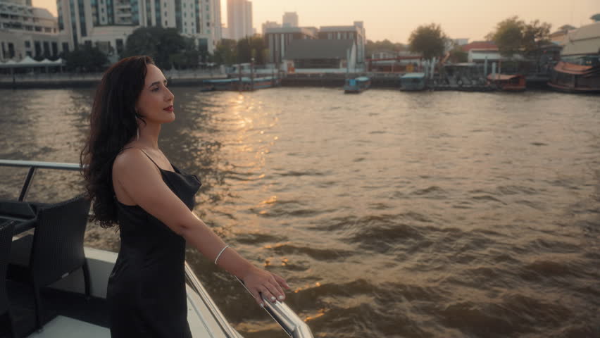 Portrait of classy Latin American woman in black cocktail gown sails on modern yacht. Happy Hispanic lady leans onto handrail looking away on luxury motorboat on vacation cruise