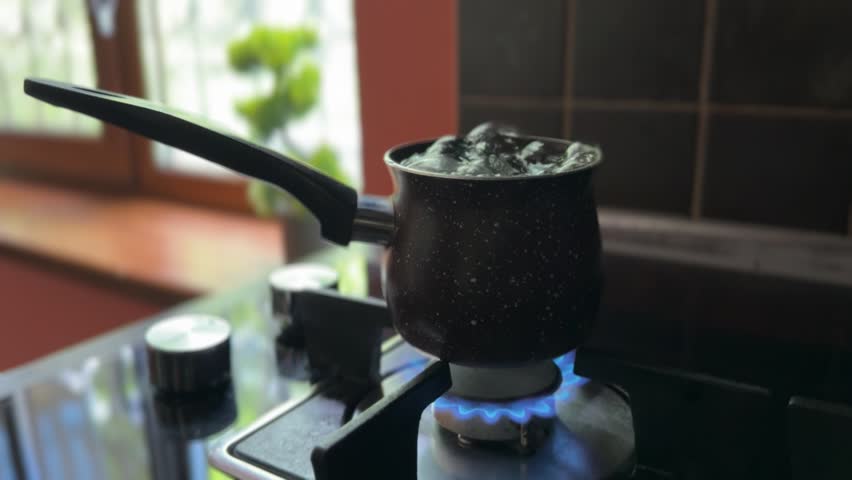 A cezve with boiling water overflows onto a lit gas burner. The bubbling water splashes on the flame, and a person quickly turns off the stove for safety.