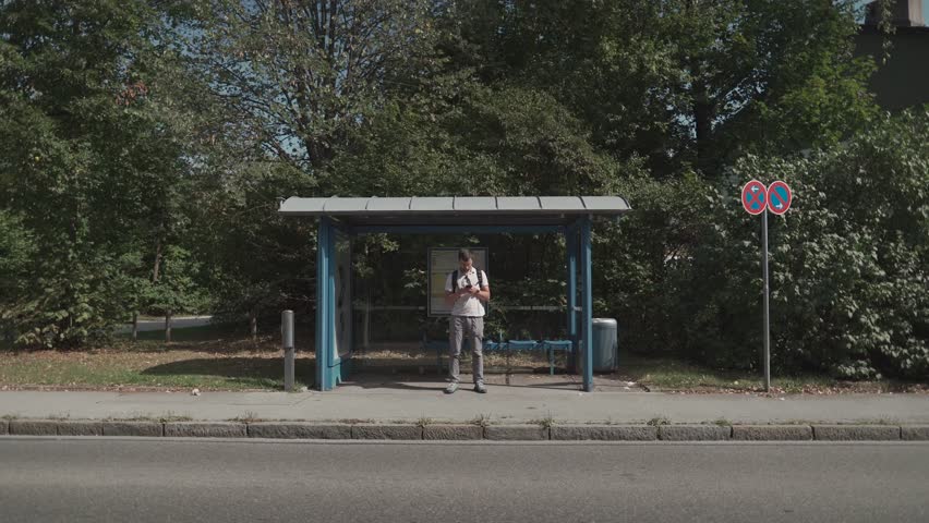 Man in Munich, Germany waits at bus stop, checking his watch. Public transport is delayed due to traffic, strike or breakdown, common urban commuting issues. Streik Verkehrsbetriebe in Deutschland. 