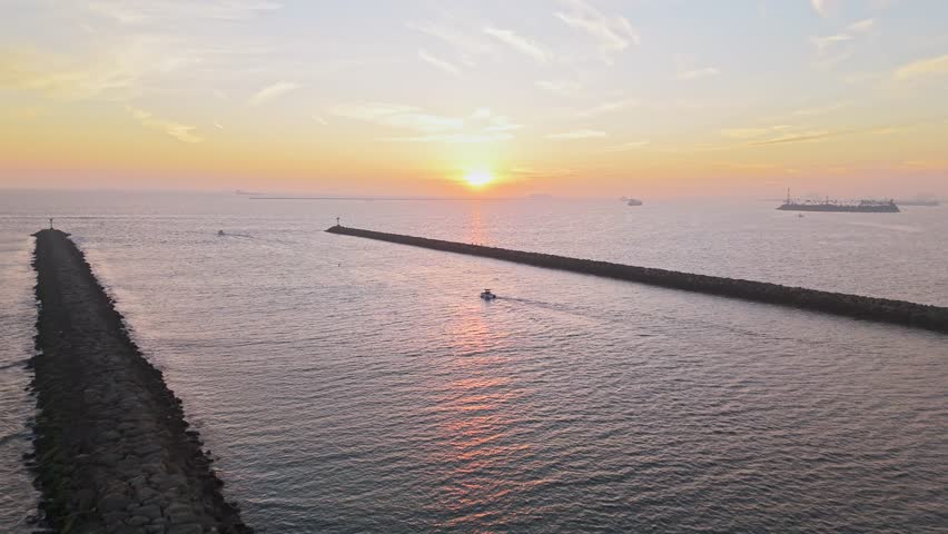 Aerial tracking shot of boat sailing out of Alamitos Bay Inlet at sunset with people fishing on a jetty, Seal Beach, California