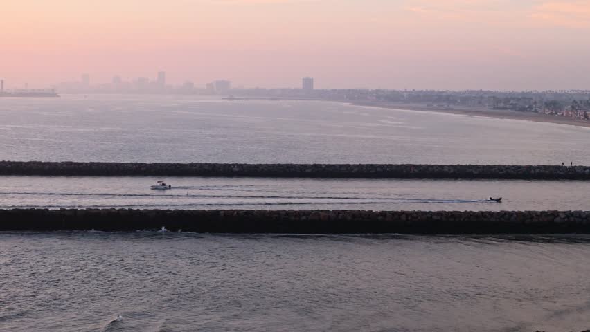 Aerial panning reveal of the sun on the horizon over Alamitos Bay Inlet with boats passing through and people fishing on the rocky jetty, Seal Beach, California