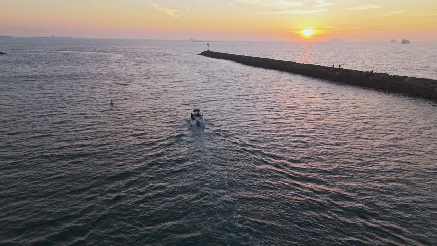 Aerial tracking shot of boat sailing out of Alamitos Bay Inlet at sunset with people fishing on a jetty, Seal Beach, California