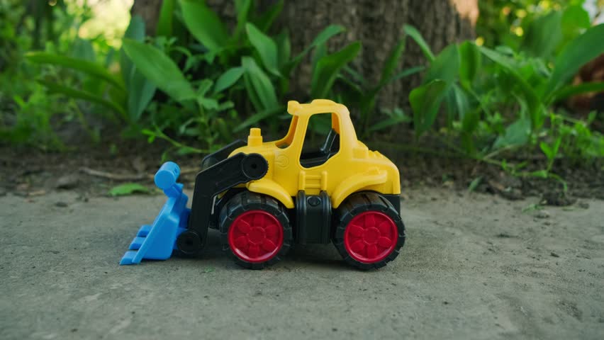 Bright Yellow Toy Construction Vehicle With Blue Scoop Resting on Dirt Near Greenery in Sunny Outdoor Setting