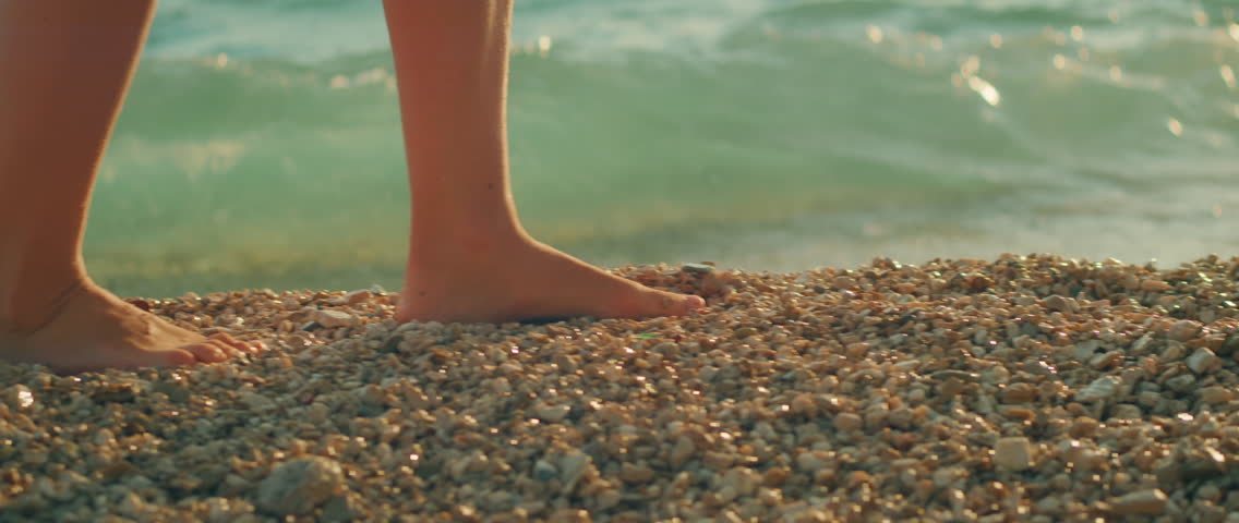 Barefoot person walking along gravel beach with gentle sea waves splashing at sunset