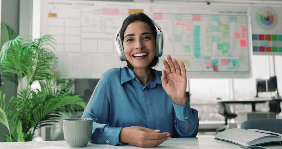 Positive young Latin business professional woman wearing headphones waving greeting hello with toothy smile at camera, talking on video conference chat, listening, nodding. Head shot portrait