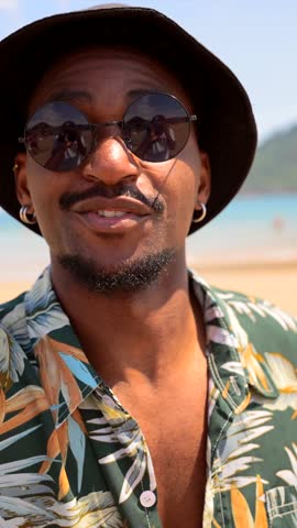 Cheerful young black man enjoying tropical beach vacation, wearing sunglasses and bucket hat, radiating pure summer joy and relaxation