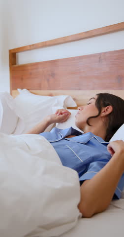 Vertical video: Woman in blue pajamas waking up in bed, reaching for alarm clock. Morning, wake-up, routine, sleepy, bedroom, lifestyle