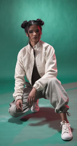 Vertical video: Young woman in casual outfit crouching confidently in stylish studio setting. Fashion, contemporary, trendy, urban, pose, attitude