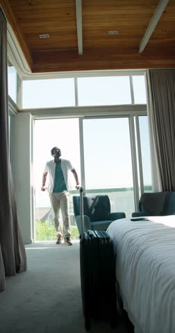 Vertical video: Man standing by window in hotel room, looking at view with suitcase nearby. Travel, tourism, leisure, accommodation, vacation, hospitality
