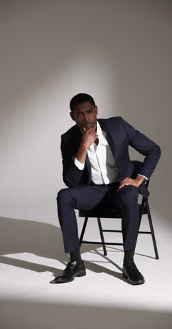 Vertical video: Confident man in stylish suit sitting on chair in modern studio setting. Fashion, elegance, menswear, formal, business, professional