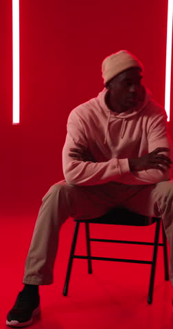 Vertical video: Young man in hoodie sitting confidently on chair in vibrant studio lighting. Fashion, confidence, modern, urban, style, male